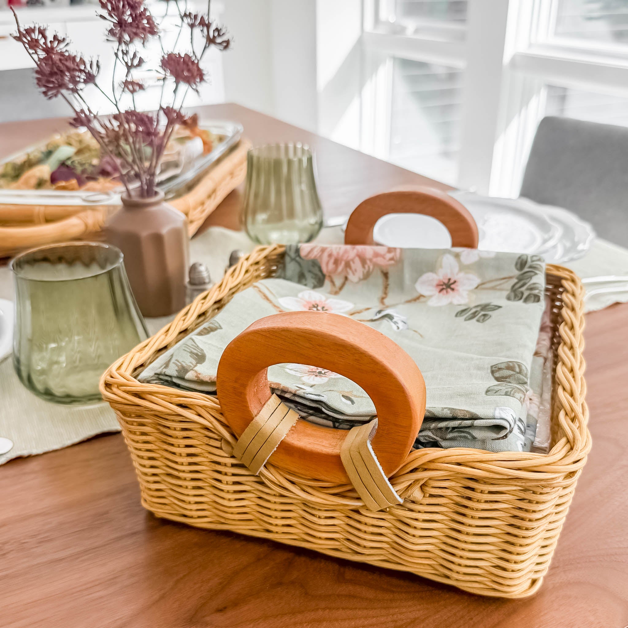 Wicker napkin basket with wooden handles on a table with a floral-patterned cloth. Wicker napkin basket in Sandstone finish, size L holds dinner-sized napkins.