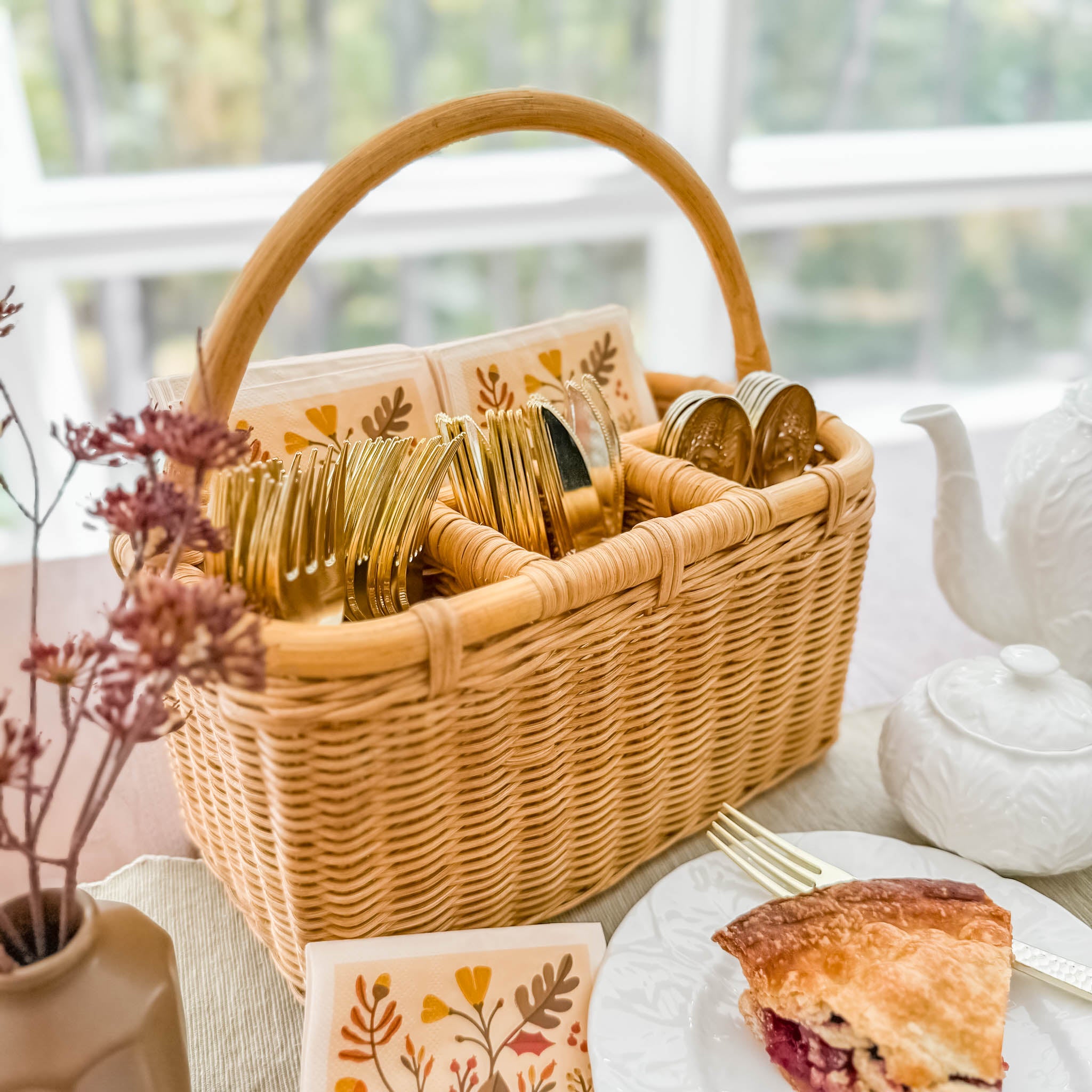 Wicker utensil caddy basket with cutlery, plates, and pie on a table. Wicker Utensil Caddy in Sandstone finish.